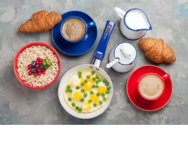 Jajecznica z groszkiem, owsianka z owocami, dwa croissanty i kawa na szarym tle.