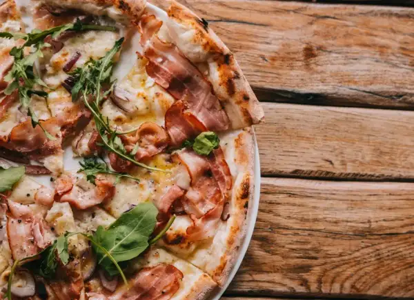 Kremowa pizza carbonara z boczkiem i rukolą na drewnianym stole.
