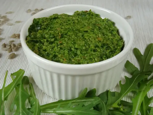 Zielone pesto z rukoli w białej miseczce, otoczone liśćmi rukoli i nasionami słonecznika.