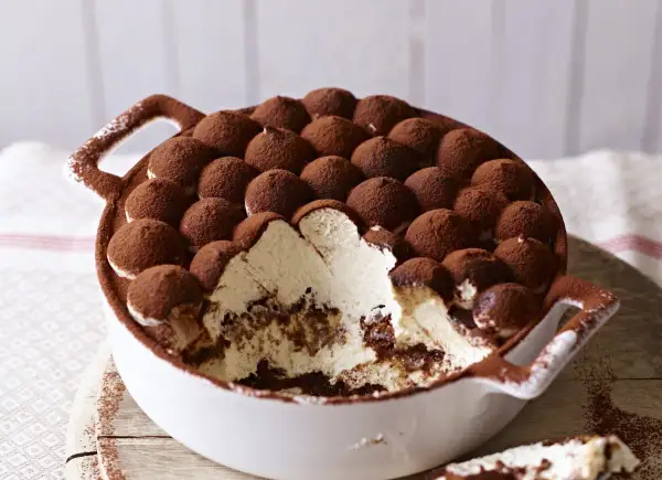 Tiramisu z mascarpone w białej misie, posypane kakao, z widocznym kremem i biszkoptami.