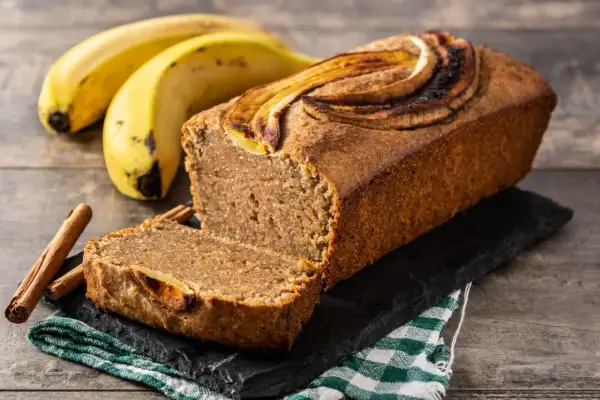 Wilgotny chlebek bananowy z plasterkami banana na wierzchu, obok banany i laski cynamonu.