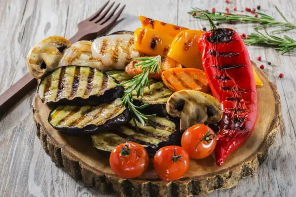 Grillowane warzywa: bakłażan, papryka, pieczarki, pomidorki i marchewka na drewnianej desce.