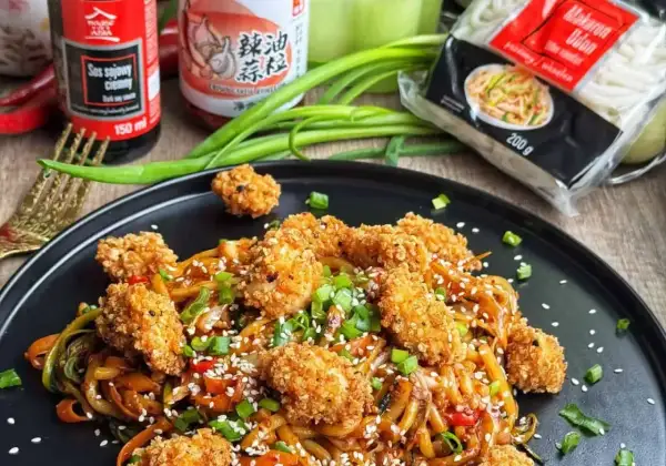 Talerz z makaronem udon, panierowanym kurczakiem, sezamem i szczypiorkiem.
