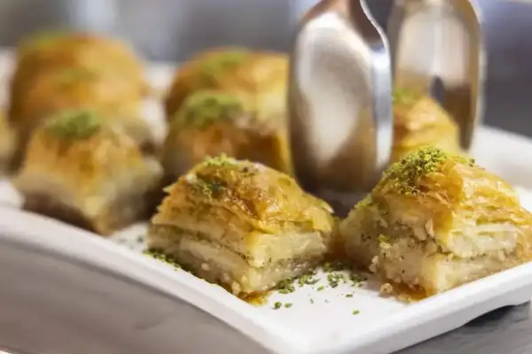 Słodka baklava z pistacjami na białej tacy, z widocznymi szczypcami do serwowania.