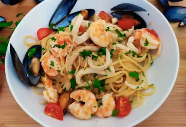 Talerz spaghetti z owocami morza: krewetkami, małżami i kalmarami, ozdobiony pomidorkami i natką pietruszki.