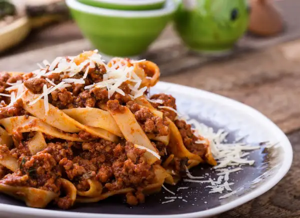 Talerz makaronu pappardelle z sosem bolognese i startym parmezanem.