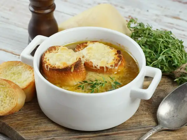Aksamitna zupa cebulowa przepis, podana w białej miseczce, z grzankami zapieczonymi z serem i gałązką tymianku. Obok kromki chleba.