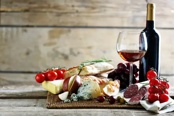 Butelka wina, kieliszek z winem, sery, pieczywo, winogrona, pomidorki i wędlina.