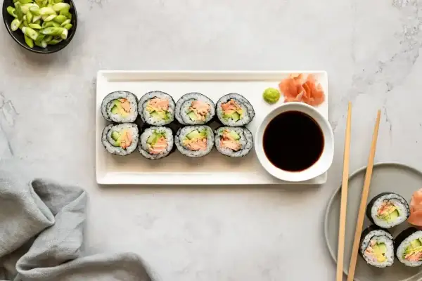 Ułożone na talerzu cienkie rolki sushi z łososiem i awokado, obok sos sojowy, wasabi i imbir.