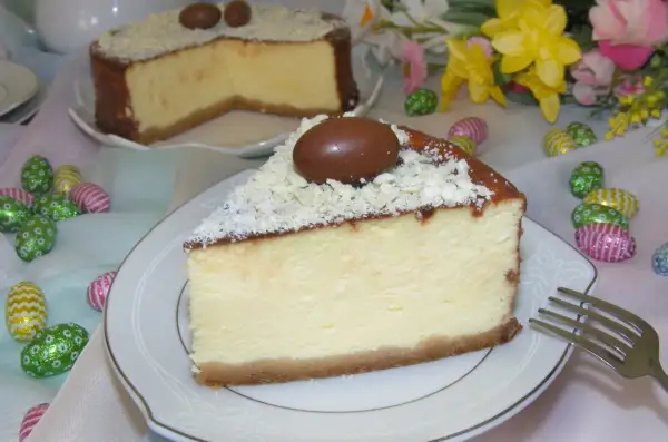 Kawałek kremowego sernika z mascarpone, ozdobiony czekoladowymi jajeczkami i wiórkami białej czekolady, na talerzyku.