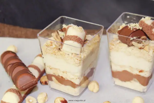 Deser Kinder Bueno w pucharkach z warstwami kremu, ciastek i czekolady, ozdobiony kawałkami batonika i orzechami.