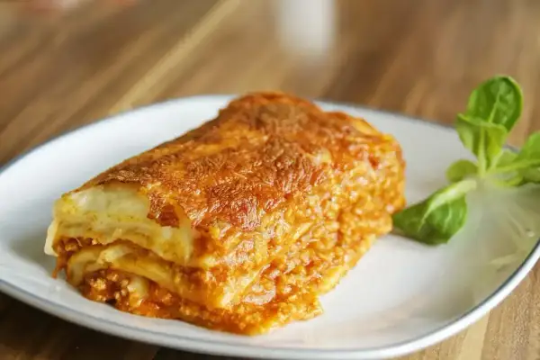 Kawałek zapieczonej lasagne z mięsem mielonym na białym talerzu, udekorowany listkami sałaty.