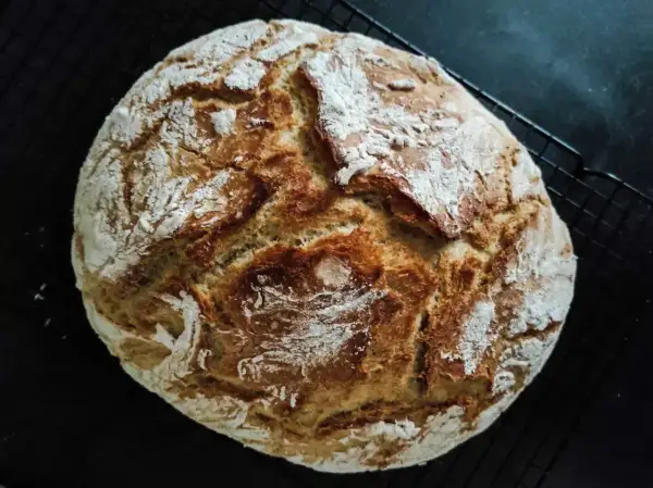 Złocisty, domowy bochenek chleba z chrupiącą skórką, posypany mąką, na czarnej kratce.