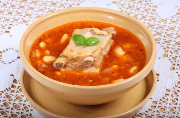 Aromaticzna zupa na żeberkach z fasolą i ziołami, podana w glinianej misce na ozdobnym obrusie.