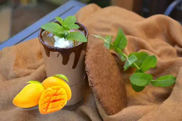 Szklanka mango lassi z czekoladą, miętą, mango i ciasteczkiem na brązowym tle.