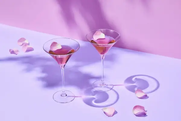 Dwa kieliszki z różowym drinkiem i płatkami róż na fioletowym tle.