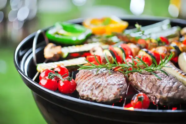 Grill z mięsem, warzywami i ziołami na tle zieleni.