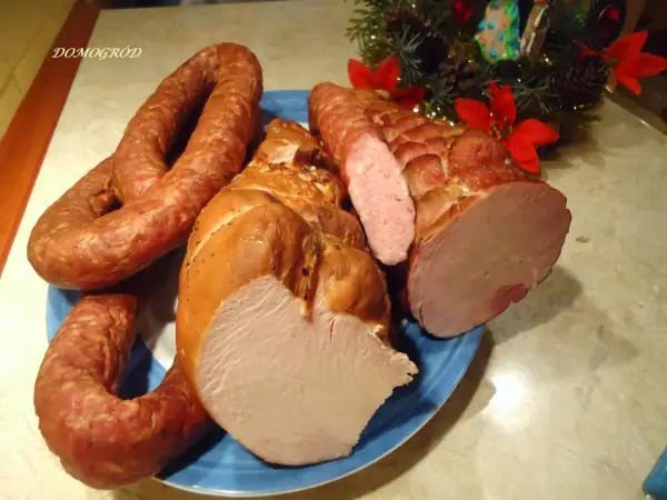 Wędzona szynka i kiełbasa na niebieskim talerzu, ozdobione gałązką choinki.