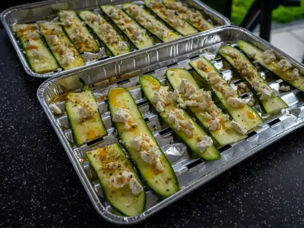 Plastry cukinii z serem feta i przyprawami na tacce aluminiowej, gotowe do grillowania.