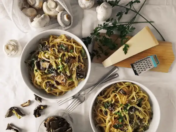 Dwa talerze makaronu z grzybami, posypane parmezanem i natką pietruszki. Obok świeże i suszone grzyby, kawałek parmezanu i tarka. Pyszny makaron z oliwą truflową.