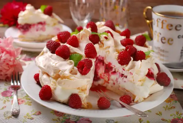 Tort bezowy z bitą śmietaną, malinami i listkami mięty na białym talerzu.
