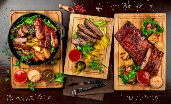 Trzy deski z grillowanym mięsem: żeberka, steki i skrzydełka kurczaka, podane z frytkami, kukurydzą i sosami.