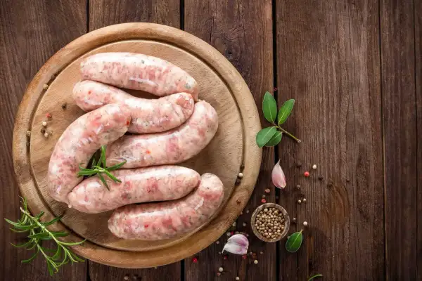 Surowa biała kiełbasa na desce, z ziołami i przyprawami. Zastanawiasz się, jak długo parzyć kiełbasę białą?