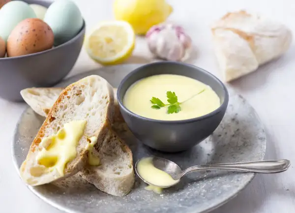 Kremowy sos aioli w miseczce, z pieczywem, jajkami, cytryną i czosnkiem w tle.