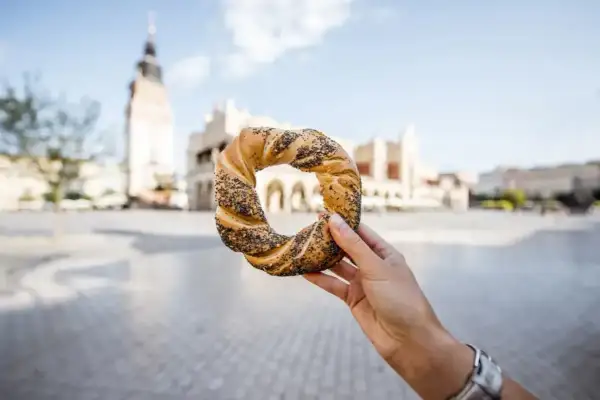 Dłoń trzyma obwarzanka krakowskiego z makiem na tle Sukiennic.
