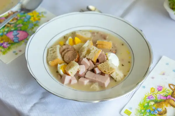 Biała zupa z kiełbasą, jajkiem i chlebem w talerzu, obok serwetki z zajączkiem.