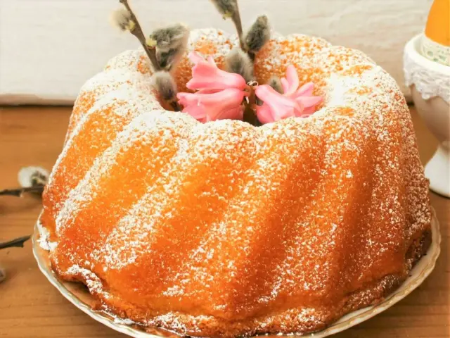 Wilgotna babka jogurtowa posypana cukrem pudrem, ozdobiona baziami i różowymi kwiatami.